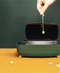 Hand placing heart necklace into Compact 40khz Ultrasonic Cleaner For Jewelry And Personal Care Devices on orange surface.