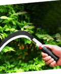 Hand holding a sickle from the Two Pack Manganese Steel Garden Sickle Tool Set, ergonomic hand weeder for yard work against greenery.