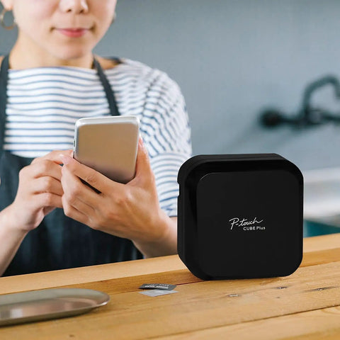 P-Touch CUBE Plus Bluetooth Label Maker PT-P710BT Portable Label Printer on table with person using smartphone