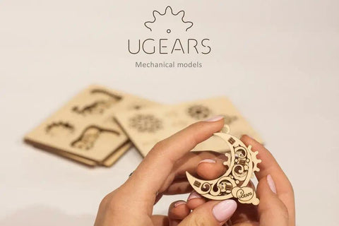 U Fidgets Gearsmas Four Mini Mechanical Puzzles Fun Hands On Self Assembly Kit In Wood Anti Stress 3D Puzzle Set Featuring Bell Snowflake Moon Snowman Plywood Building Toys For Adults And Kids Ages 5 Plus wooden moon puzzle held in hands, with assembled mini parts and flat sheets in background
