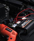 Ride Ready 500W Car Power Inverter Converts 12V DC To 110V AC With Dual USB Ports And Slick Compact Build To Keep All Your Devices Juiced Up While You Cruise On The Open Road, mounted on a car battery with red cables.