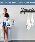 Woman in laundry room near black metal towel rack with 'Towels' sign, holding laundry basket; laundry decor setup idea.