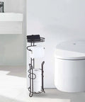 Heavyweight Free Standing Toilet Paper Roll Holder With Reserve Shelf Bronze Finish next to modern toilet and sink.