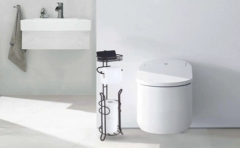 Heavyweight Free Standing Toilet Paper Roll Holder With Reserve Shelf Bronze Finish next to modern toilet and sink.