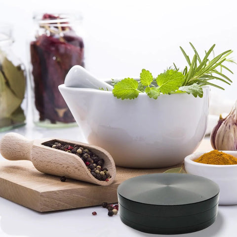Front view of a 4-inch black herb and spice grinder on a kitchen counter with mortar and pestle and spices
