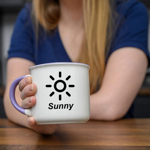 Person holding a white mug with purple handle featuring a sun design and the word 'Sunny'.