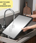 Front view of a large titanium cutting board with black border being rinsed under a kitchen faucet