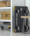 Heavy Duty Folding Push Cart With Reinforced Platform stored upright next to shelves, showcasing swivel wheels and grey plastic plate.
