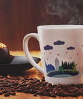 White ceramic mug with a colorful landscape print; steam rises from the cup on a wooden table with scattered coffee beans.