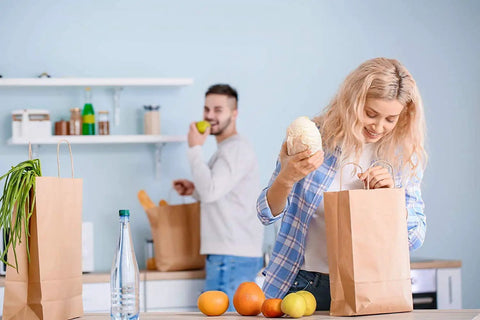Woman packing groceries in kitchen with Bulk 500 Pack Heavy Duty Kraft Paper Bags 10x13 Inches With Handles, man in background.