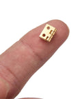 Close-up of a single mini hinge on fingertip, part of a 100 Pack Mini Hinges With 400 Replacement Screws In Gold Copper Finish Four Hole Butt Hinges 10x8mm Ideal For Jewelry Cases Cabinets Closets Dollhouses Mirrors And Picture Frames Easy Install.