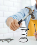 Man using a hammer to install a black steel coil spring from the Heavy Duty Six Inch Sofa Spring Repair Kit.