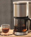 Drip Coffee Maker 1250W Power, 1.25L glass carafe, stainless steel filter, anti-drip design, beside a filled glass cup.