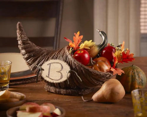 Cornucopia centerpiece with fall fruits and leaves, wicker basket marked with 'B' on a wooden table, surrounded by autumn decor.