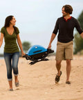 Two people carrying Q1200 Portable Propane Grill For Travel And Camping Blue With 1 Burner Cast Iron Grates Electronic Ignition 8500 BTU on beach.