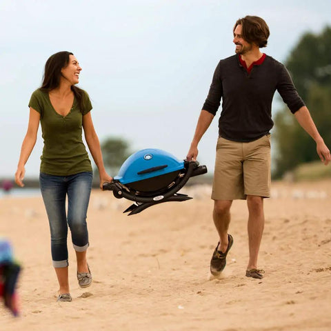 Two people carrying Q1200 Portable Propane Grill For Travel And Camping Blue With 1 Burner Cast Iron Grates Electronic Ignition 8500 BTU on beach.