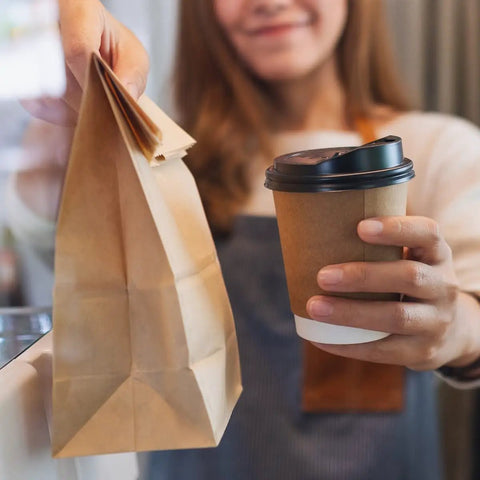 Person holding 100 Pack Brown Kraft Paper Lunch Bags With 4lb Capacity, 5 By 2.95 By 9.45 Inches Fold Top Disposable Bags alongside coffee cup.