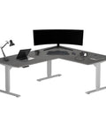 L Shaped Electric Standing Desk With Effortless Height Adjustment Serving As A Rock Solid Corner Workspace Measuring 72 By 60 Inches Straight Up Crafted For Daily Use Boasting Heavy Duty Gray Frame Paired With A Top Shelf Gray Oak Desktop featuring laptop and monitor.