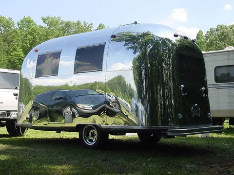 Shiny chrome vintage trailer reflecting surrounding greenery and vehicles, parked on grass, showcasing its sleek design.