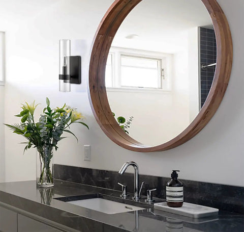 Modern Farmhouse Glass Cylinder Wall Sconce 1 Light Black Vanity Light Fixture mounted in a bathroom with round mirror and flowers.