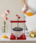 Heavy Duty 1.6 Gallon Stainless Steel Manual Press For Wine Cider Honey And Juicing on a table with fresh fruit and flowers.