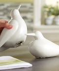Person placing a white ceramic bird on a table next to another bird, with a towel nearby in a kitchen setting.