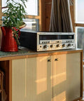 Vintage stereo on cabinet with brass knobs, next to a red watering can and green plant in a sunlit room.