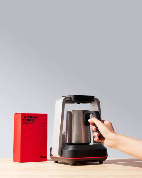 Person using Turkish Coffee Maker Electric with Open Form Design, SandBrew Technology, stainless steel, beside red coffee packaging.