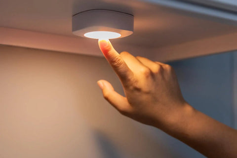 A hand tapping to turn on a Tap On Off LED Light Stick-On Battery Powered Wireless Portable Light For Cabinets and Closets No Fuss White Under Cabinet Lighting Pantries Attics Bathrooms RVs Requires 3 AA Batteries 2 Pack mounted under a white surface.