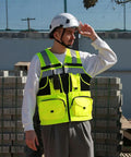 High Visibility Reflective Safety Vest With Heavy Duty Construction And Plenty Of Pocket Space For Tools On Any Job Site Hella Bright Orange Unisex Work Vest In Three X Large Size worn by worker with helmet in construction area.