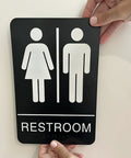 Person installing unisex braille restroom sign with graphical symbols and 3M double sided tape, 9 by 6 inches, ADA compliant.
