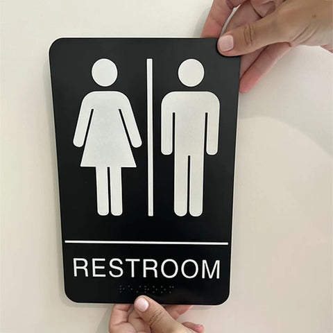 Person installing unisex braille restroom sign with graphical symbols and 3M double sided tape, 9 by 6 inches, ADA compliant.