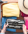 Hands holding Go-To Vintage Airplane Travel Toiletry Bag for Women and Men over open suitcase with folded clothes and hat.