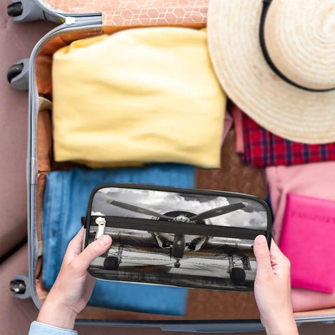 Hands holding Go-To Vintage Airplane Travel Toiletry Bag for Women and Men over open suitcase with folded clothes and hat.
