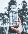 Person holding 1-liter soda bottle outdoors, part of 3 Pack Reusable 1 Liter Sparkling Water Bottles For Soda Makers With Hermetically Sealed Caps Keep Carbonation Longer Includes Gift Kidscare 14 Inch Bottle Cleaning Brush For Easy Cleaning bundle.