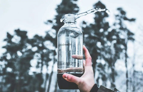 Person holding 1-liter soda bottle outdoors, part of 3 Pack Reusable 1 Liter Sparkling Water Bottles For Soda Makers With Hermetically Sealed Caps Keep Carbonation Longer Includes Gift Kidscare 14 Inch Bottle Cleaning Brush For Easy Cleaning bundle.
