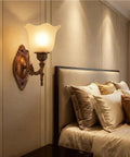 Antique Copper Wall Sconce Light European Vintage Design with Flower Glass Shade Carved Base Indoor Wall Lamp for Bedroom Living Room Hallway Bedside Decor E26/27 next to a bed with plush pillows.