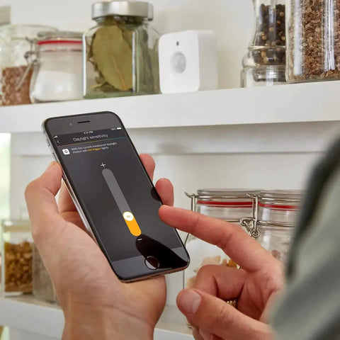 Person adjusting Indoor Smart Motion Sensor 2 Pack with Daylight Sensing settings on smartphone in kitchen.