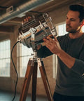 Man adjusting Nautical Style Chrome Spotlight Floor Lamp With Brown Wood Tripod Stand, rustic charm in industrial setting.