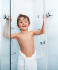 Child standing in shower with clear glass doors and white towel, demonstrating frameless shower design