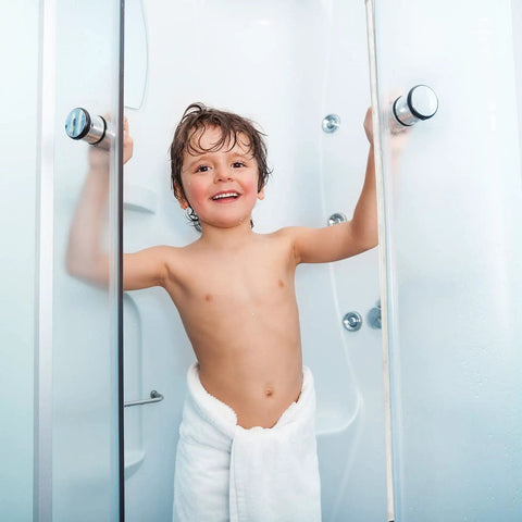 Child standing in shower with clear glass doors and white towel, demonstrating frameless shower design