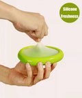 Hand stretching green silicone top, part of Set Of Four Reusable Fruit And Veggie Savers For Keeping Avocados Tomatoes Apples Garlic And Onions Fresh Longer In The Fridge Space Saving Design Makes Kitchen Organization Easy