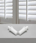 Two white spring loaded shutter pins for plantation window shades on a gray surface with window blinds in the background.