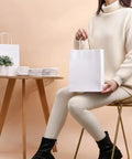 Woman holding White Kraft Paper Gift Bags With Handles Pack Of 25 Medium 8 By 4.5 By 10 Inches Tough Durable Recyclable Shopping Bags