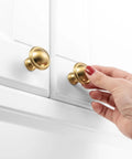 Hand holding brushed gold cabinet knob on white cabinet, part of 10 Pack One And A Quarter Inch Brushed Gold Cabinet Knobs For Cabinets Drawers Dressers Cupboards Sturdy Solid Metal Hardware Built To Last In Humid Kitchens And Bathrooms