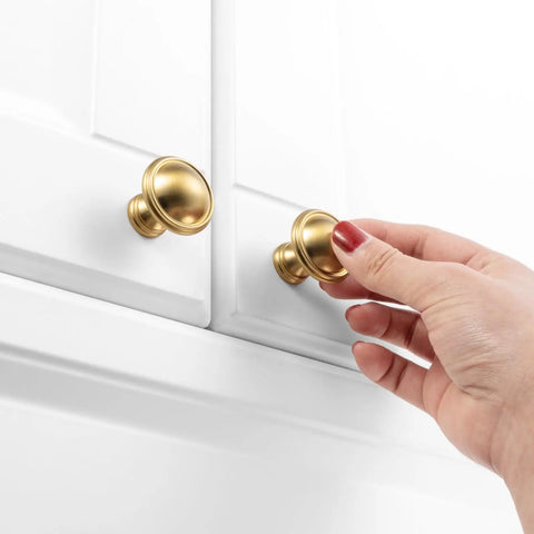 Hand holding brushed gold cabinet knob on white cabinet, part of 10 Pack One And A Quarter Inch Brushed Gold Cabinet Knobs For Cabinets Drawers Dressers Cupboards Sturdy Solid Metal Hardware Built To Last In Humid Kitchens And Bathrooms