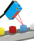 Compact Diffuse Photoelectric Sensor With Background Suppression And 2mm Red Laser, No Fuss Install, Sensing Range 20 To 300 Mm, 10 To 30 VDC, PNP Output, Unaffected By Background Color, Ideal For Transparent Objects. Blue compact sensor over conveyor detecting colored cylindrical parts.