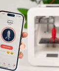 Smartphone showing rocket print on app, Beginner Friendly 3D Printer For Kids With Easy Setup And Safe Design in the background