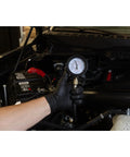Mityvac MV5530 engine compression test gauge held by gloved hand under a car hood, hose connected to engine