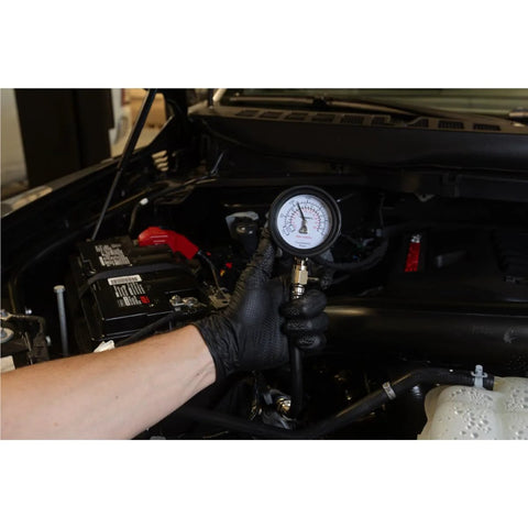 Mityvac MV5530 engine compression test gauge held by gloved hand under a car hood, hose connected to engine